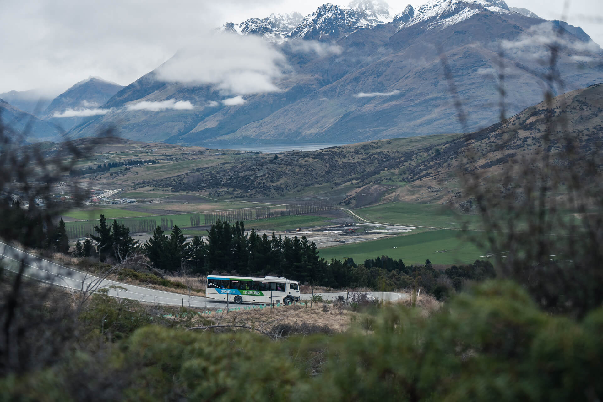 Queenstown Ski bus driving on a winding mountain road surrounded by trees, overlooking a vast valley with green fields, a lake, and snow-capped mountains partially covered by clouds in the background.