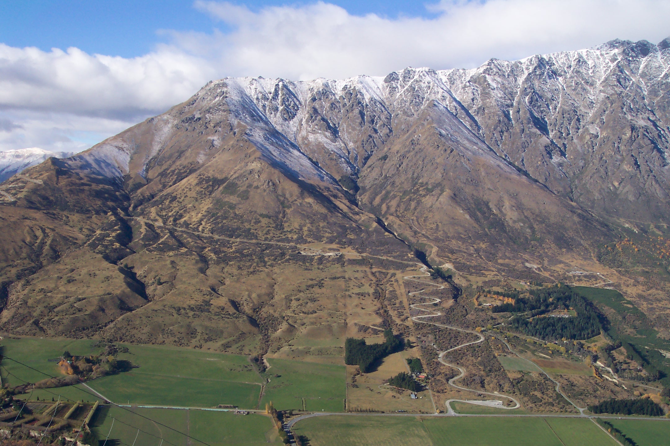 Aerial view of a mountain range with snow-dusted peaks and a winding road leading up from green fields below.